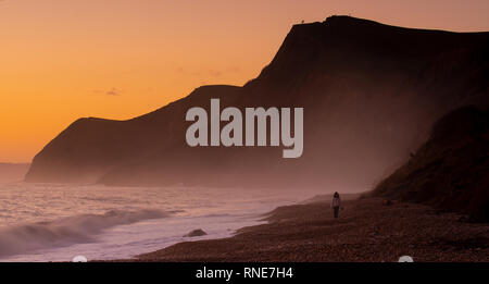 Eype, Dorset, Großbritannien. Februar 2019. UK Wetter: Eine Frau geht am Strand von Eype entlang mit Thorncombe Beacon in der Ferne, während der Himmel bei Sonnenuntergang orange über der Jurassic Coast glüht. Quelle: DWR/Alamy Live News Stockfoto