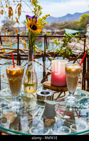 Zwei Ice Coffee Gläser mit rotem Strohhalm, Vase mit Blumen, Aschenbecher und rote Kerze auf dem Metall Glas Tisch von exotischen Pflanzen umgeben. Romant Stockfoto