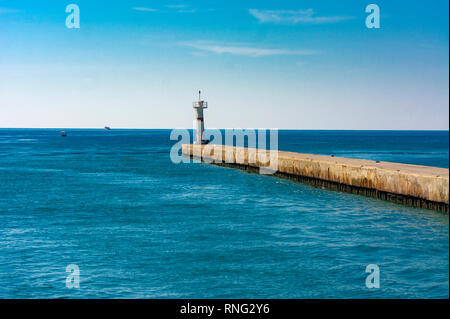 Schöne Aussicht auf den Leuchtturm durch eine lange Betonpfeiler gegen den blauen Himmel und das blaue Meer. Beleuchtung der Reisenden. Stockfoto