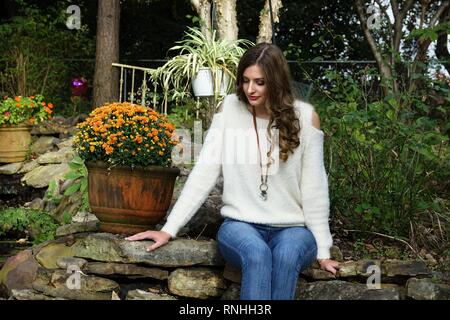 Junges Land Frau oder ein Mädchen mit langem Haar trägt einen Pullover und Jeans in einer natürlichen Umgebung mit Blumen und eine Felswand Stockfoto