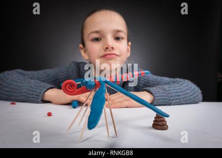 Weitwinkel- Portrait von ein junges Mädchen 9-12 Jahre alt im Defokussierten Form, im Vordergrund auf einem weißen Tisch steht eine Libelle blau-rot pastelina, w Stockfoto