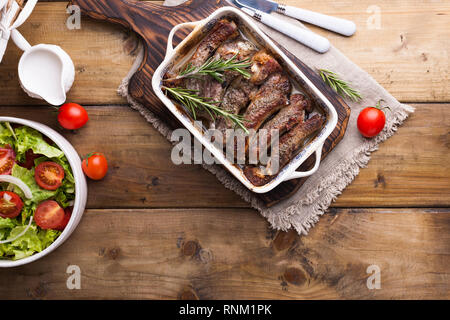 Gegrillte Schweineribs. Fleisch mit Gewürzen und Kräutern in Gerichten auf Holzgrund. Mittagessen Barbecue mit Salat und Sauce. Freier Speicherplatz für den Text Stockfoto