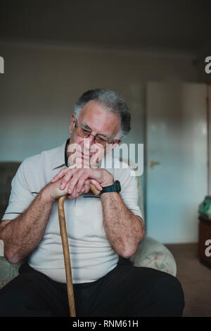 Älterer Mann ist traurig mit seinen Kopf auf seinen Spazierstock im Wohnzimmer seines Hauses. Stockfoto