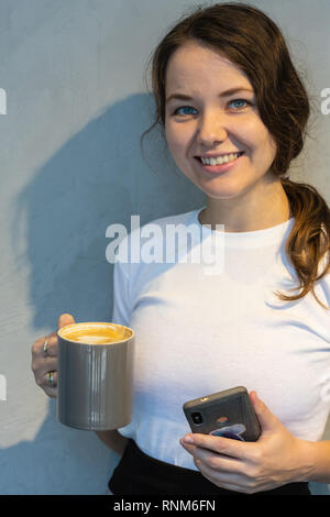 Gut aussehende junge Hipster girl in stilvoller Kleidung und Schattierungen Nachrichten online und Kontrollen Newsfeed auf Smart Phone, sitzt im Café allein, schaut heraus Stockfoto