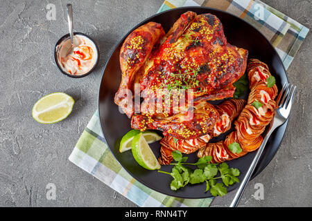 Gebratenes Huhn mit Tomatensauce und ganze Senf erstickt serviert mit hasselback süsse Kartoffel und saure Sahne sriracha Soße auf einer schwarzen Platte Stockfoto