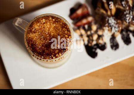 Makro Foto Tasse heißen Latte mit caramel Kruste gebacken und Brötchen mit Banane und Erdbeere auf dem hölzernen Tisch in Coffe-sho. Kaffee Konzept. T Stockfoto