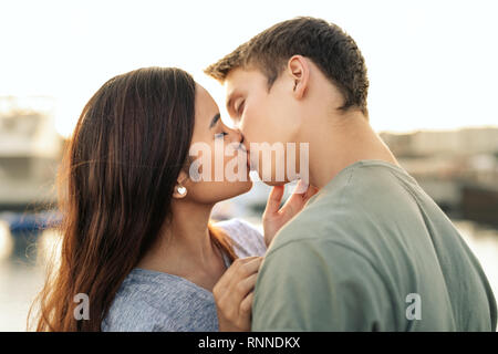 Liebevolle junge Paar teilen einen Kuss im Stehen, Arm in Arm durch einen Hafen am späten Nachmittag Stockfoto