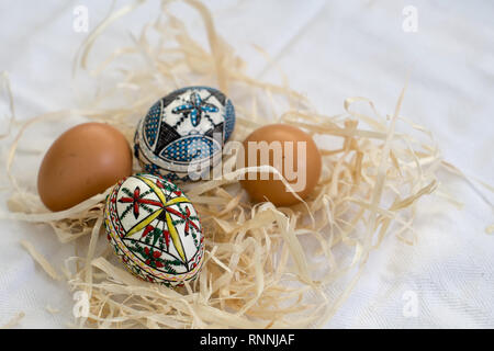 Traditionelle handgefertigte Ostereier im Stroh auf einem weißen Tuch und wenige Organische frische Eier im Hintergrund. Traditionelle Muster von Bucovina Stockfoto