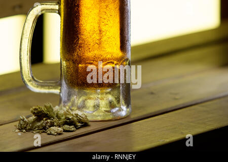 Isolierte gefrorenen Bierkrug auf Holz Tisch, getrocknete Hopfen Blätter Stockfoto