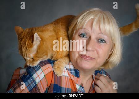 Schön gealterte Frau umarmte ihre rote Katze auf grauem Hintergrund. Stockfoto