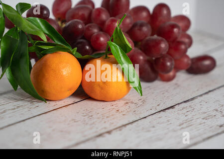 Zwei goldene orange Mandarinen mit grünen Blättern hinter sitzen auf einem rustikalen weiß Holz Tisch Hintergrund mit roten Trauben befestigt Stockfoto