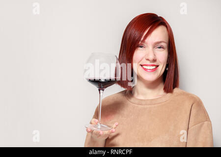 Junge schöne rothaarige Mädchen Sommelier mit Hellen Lippenstift auf weißen Hintergrund ist und lächelnd Glas Rotwein an professionelle blinde Tasten Stockfoto