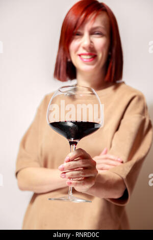 Junge schöne rothaarige Mädchen Sommelier mit Hellen Lippenstift auf weißen Hintergrund ist und lächelnd Glas Rotwein an professionelle blinde Tasten Stockfoto