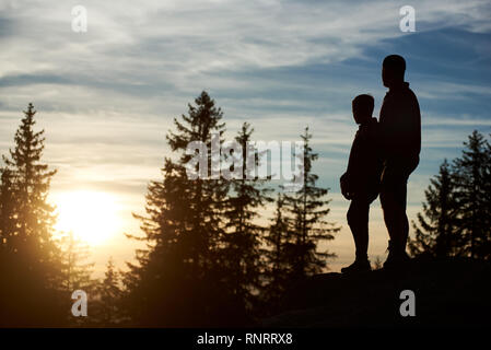 Silhouetten der junge Vater und Sohn stehen auf dem Gipfel des Berges in den Abend. Mann und Kind genießen Sie einen wunderschönen Sonnenuntergang, Bäume auf verschwommenen Hintergrund. Stockfoto