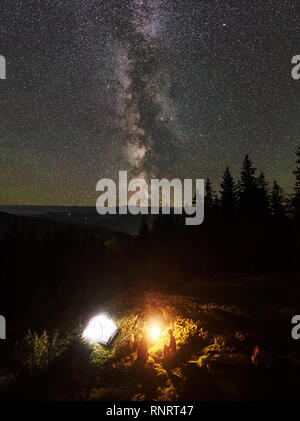 Vertikaler nacht Sommer Camping in den Bergen. Blick von oben auf die paar Wanderer zusammen neben Feuer und glühende touristische Zelt unter schöne Nacht Sternenhimmel voller Sterne und Milchstraße sitzt. Stockfoto