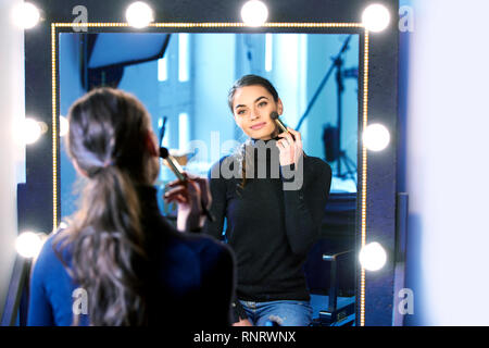 Portrait von wunderschöne junge Frau ihr Make-up auftragen beim Sitzen am Spiegel im Fotostudio. Stockfoto
