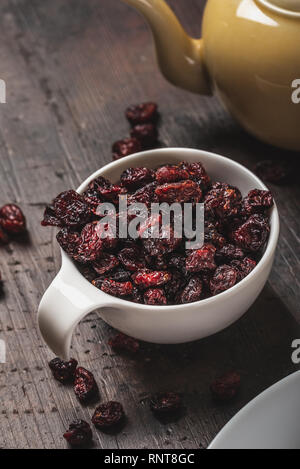 Vertikale Foto der weißen Schale voller getrocknete Cranberries. Die Moosbeere dunkelrote Farbe und sind um verschüttet. Cup ist auf vintage Holzbrett w platziert Stockfoto