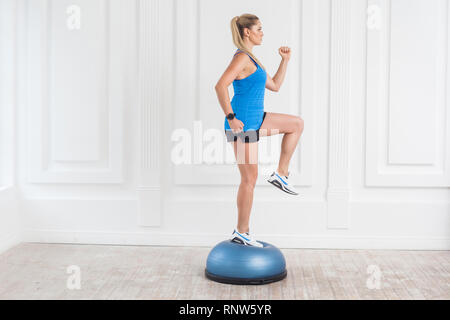 Seitenansicht Portrait von Konzentration sportlich schönen jungen athletischen blonde Frau in schwarzen Shorts und Blue Top arbeiten im Fitnessstudio tun exersice in Bosu ba Stockfoto