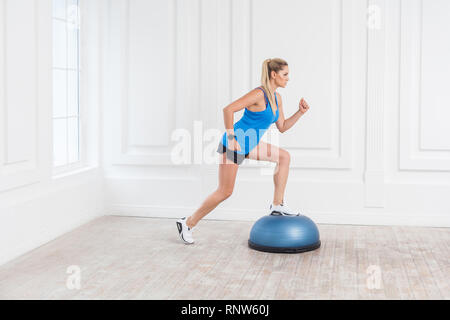 Seitenansicht Porträt der fokussierten sportliche junge athletische blonde Frau in schwarzen Shorts und Blue Top arbeiten im Fitnessstudio tun exersice in Bosu Balance Trainer, m Stockfoto