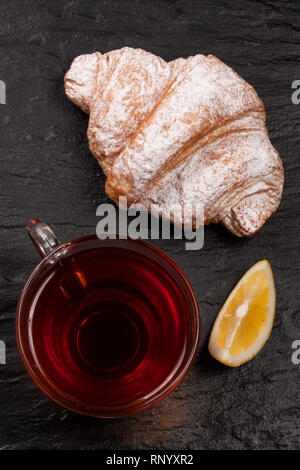 Croissant mit einer Tasse Tee auf einem schwarzen Stein. Ansicht von oben. Stockfoto