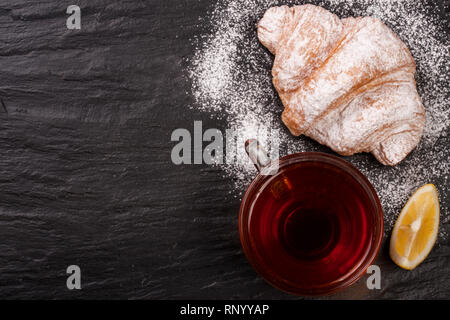 Croissant mit einer Tasse Tee auf einem schwarzen Stein Hintergrund mit Kopie Platz für Ihren Text. Ansicht von oben. Stockfoto