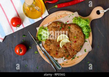 Große Wiener Schnitzel auf einem Holzbrett mit Zitrone auf einem dunklen Hintergrund. Fleischgericht. Ansicht von oben, flach. Stockfoto