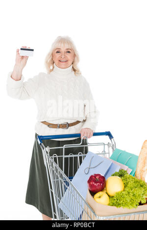 Freundliche ältere Frau mit Kreditkarte in der Nähe von Shopping Trolley isoliert auf weißem Stockfoto