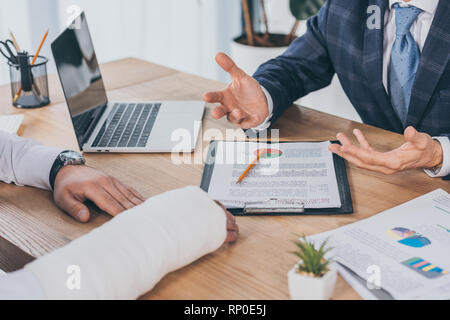 7/8-Ansicht der Geschaeftsmann in der blauen Jacke Standortwahl am Tisch gegenüber Arbeiter mit gebrochenen Arm in Büro, Entschädigung Konzept Stockfoto