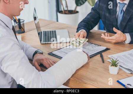 7/8-Ansicht der Geschaeftsmann in der blauen Jacke am Tisch sitzen und Geld zu geben Arbeiter mit gebrochenen Arm in Büro, Entschädigung Konzept Stockfoto