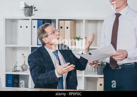 Im mittleren Alter unzufrieden Geschäftsmann am Tisch sitzt und spricht mit Arbeiter in der Nähe von mit Dokumenten in Office, Entschädigung Konzept Stockfoto