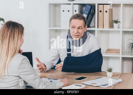 Im mittleren Alter Arbeiter in Hals mit gebrochenen Arm sitzen am Tisch und reden blonde Frau im Büro, Entschädigung Konzept Stockfoto