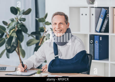 Arbeiter in Neck Brace und arm Bandagen am Tisch sitzen, lächeln und Schreiben mit Kugelschreiber im Büro, Entschädigung Konzept Stockfoto