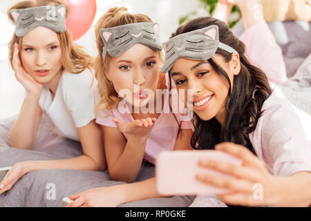 Schönen Lächeln multikulturelle Mädchen in schlafmasken im Bett lag und unter selfie auf Smartphone während Pyjamaparty Stockfoto