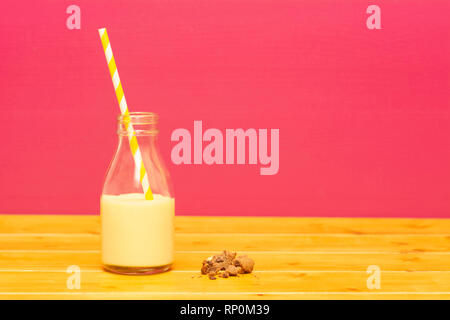 Ein Drittel pint Glas Milch Flasche halb voll mit Banane Milchshake mit retro Stroh und ein halbes gegessen Chocolate Chip Cookie, auf einen hölzernen Tisch gegen Stockfoto