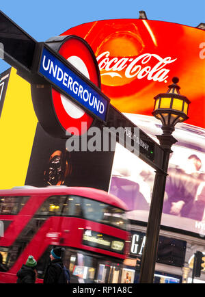 London Piccadilly Circus mit traditioneller U-Bahn-U-Bahn-U-Bahn-Schild und Coca Cola-Neon-Banner für digitale Plakatwerbung hinter und verwischter roter, emissionsarmiges Hybrid-London Bus in der Abenddämmerung Stockfoto