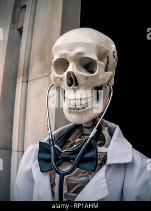 DR DEATH CONCEPT MEDICAL NHS WARTELISTE Schädelskelskelett lächelnd gekleidet in einen weißen Mantel und eine Fliege mit Stethoskop, das als „Doktor Tod“ fungiert. Eine humorvolle Erinnerung außerhalb einer medizinischen Einrichtung, gesund zu bleiben Stockfoto