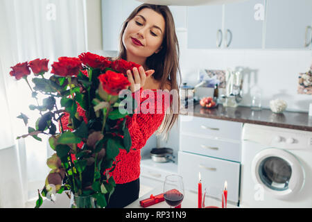 Junge Frau gefunden Blumenstrauß aus Rosen mit Kerze, Wein und Geschenkbox auf Küche. Happy girl duftende Blumen. Valentines Tag Überraschung Stockfoto