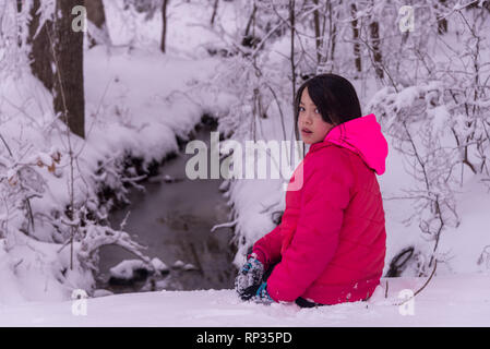 Junge asiatische Mädchen sitzen im Schnee vor Stream Stockfoto