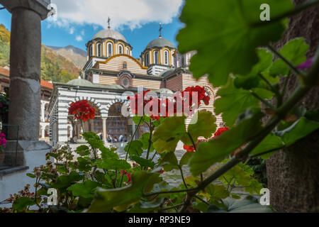 Die Kirche von Kloster Rila, Bulgarien unscharf im Hintergrund hinter hellen roten Geranien um innere Kante von seinem Hof platziert Stockfoto