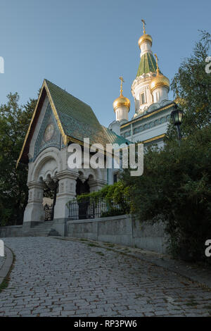 Gepflasterten Straße führt zu schönen St. Nikolas Russische Kirche, Sofia, Bulgarien. Am späten Nachmittag Sonne Highlights drei goldene 'Zwiebel' Kuppeln und Verkleidung Stockfoto