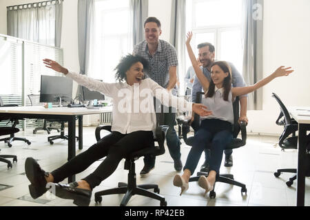 Junge fröhliche multirassischen diverse Kollegen Spaß reiten auf Bürostühlen in Coworking Space aufgeregt. Aktive glücklich tausendjährigen Arbeiter ein br Stockfoto