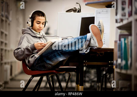 Teenager mit seinen Füßen zu sitzen und ein Buch lesen mit Kopfhörern in einer Bibliothek. Stockfoto