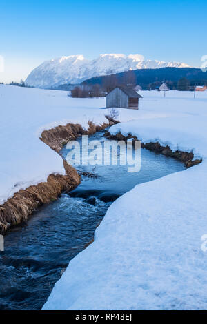 Kleine gekrümmte Creek River mit Büschen auf Bank schlängelt sich durch ein schneebedecktes Feld mit Berg Grimming, Steiermark, Österreich Stockfoto