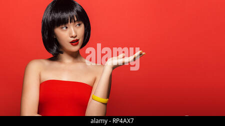 Porträt der attraktive junge asiatische Frau mit kurzen Haaren bei copy Raum zeigt. Stilvolle weibliche Modell zeigen Sie ein Produkt auf roten Hintergrund. Stockfoto