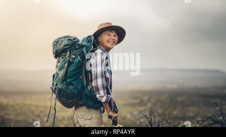 Seitenansicht eines älteren Frau Rucksack tragen auf einem Hügel beim Trekking. Lächelnd senior Frau während einer Wanderung Kampagne aufgeregt. Stockfoto