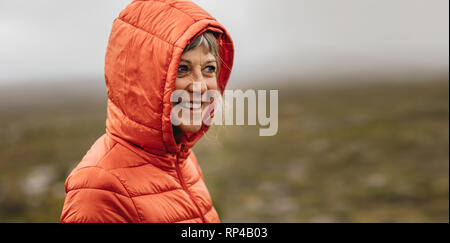 Lächelnde Frau in eine warme Kapuzenjacke stand im Freien. Freundliche ältere Frau, die auf einem Hügel entfernt. Stockfoto
