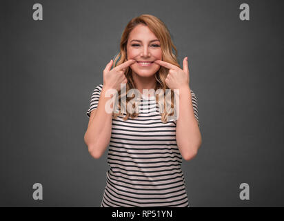 Eine junge Frau ihren Mund in ein unechtes Lächeln vor grauem Hintergrund. Fake Lächeln. Stockfoto