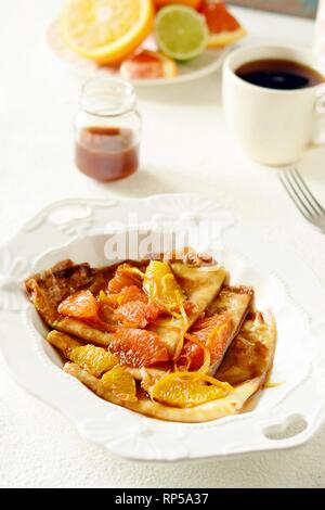 Pfannkuchen Crepe Suzette zum Frühstück mit orange Karamell-sauce, Orangenscheiben, Kalk und Orangenschale und eine Tasse Kaffee. Dessert französische Küche. Sunda Stockfoto