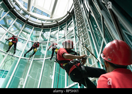 Arbeiter in Sicherheitsausrüstung reinigen Hochhausfenster in einem Glasgebäude, indem sie Seile und Gurte verwenden, um schwierige Stellen zu erreichen und Teamarbeit zu zeigen Stockfoto