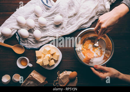 Blick von oben Frau Hände mischen Saure Sahne, Zucker und Eigelb in Metall Schüssel Neben Eiern, gewürfelte Butter und Mehl auf braune Holztisch. Prozess der Stockfoto